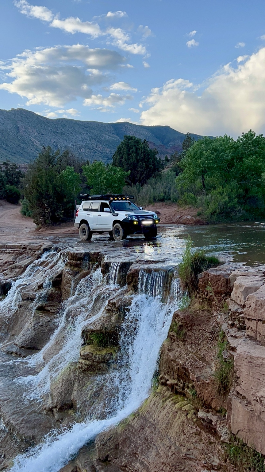 White overland vehicle crossing a shallow river above a waterfall